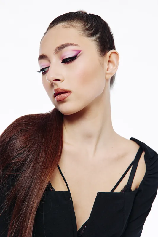 Portrait of a young woman with creative pink eyeshadow and glossy lips wearing a black top with strap details. Her long hair is pulled back for a striking beauty and fashion look.