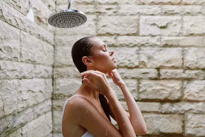 woman showering outdoors under rain showerhead with wet hair, natural stone wall background, skincare, relaxation, self-care moment in warm daylight