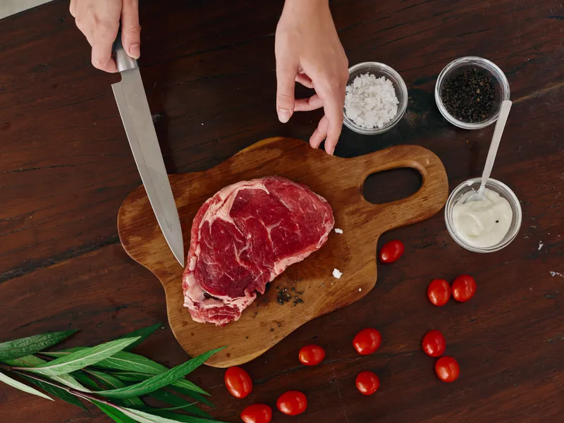 Женщина с ножом в руке нарезает свежее стейковое мясо для жарки на кухне, на столе соль, перец и другие специи, красные помидоры черри и зелень, приготовление ужина в домашней кухне
