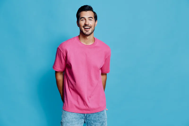 Smiling young man wearing a pink t-shirt, standing with hands behind back on a bright blue background. Cheerful and relaxed vibe, capturing youthful energy.