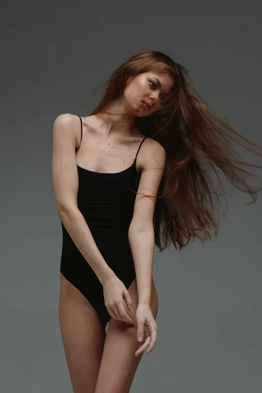 Slender woman with long hair in black bodysuit posing with movement against gray background
