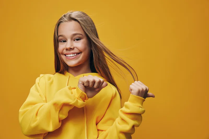 Smiling girl with long hair wearing yellow hoodie posing on yellow background, happy child with casual clothes and cheerful expression looking at camera.