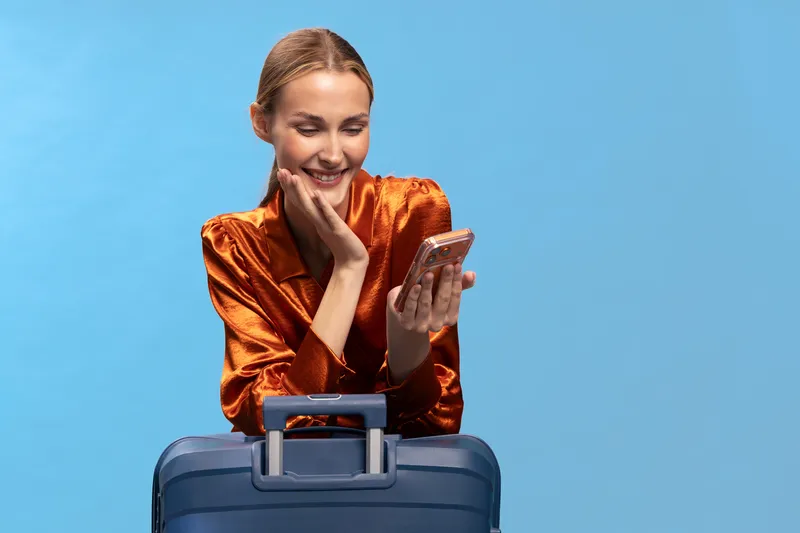 Smiling woman in shiny orange blouse looks at smartphone while leaning on blue suitcase. Studio portrait with bright blue background. Concept of travel, communication, and technology use.