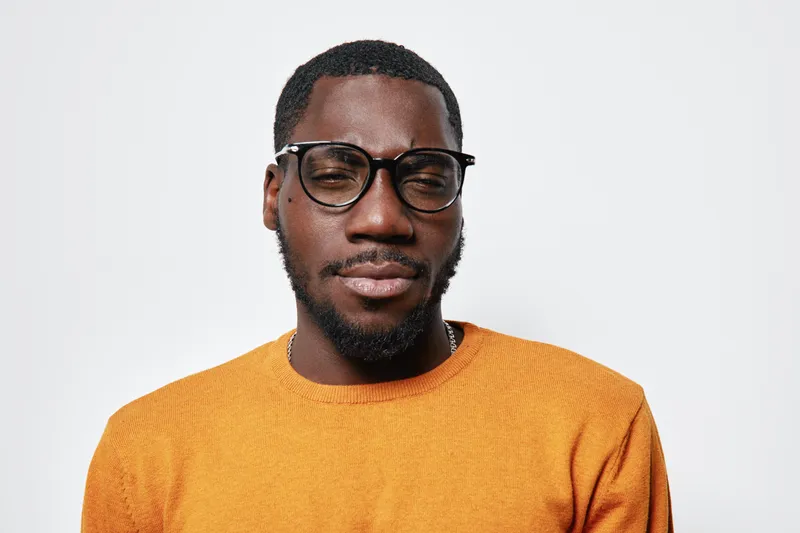 Portrait of African American man with dark skin wearing glasses and orange sweater against white background. Confident expression, casual style and studio lighting for modern look.