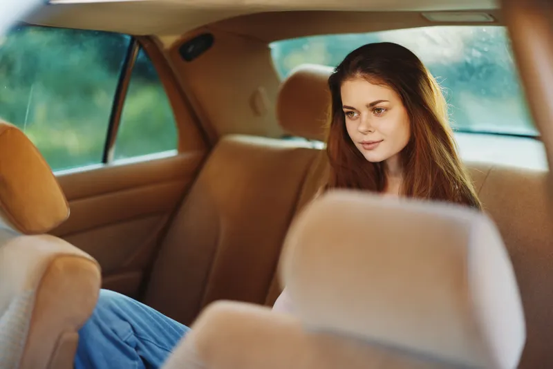 A pensive young woman with long brown hair sitting in the backseat of a car, reflecting thoughtfully against a blurred natural background, embodying tranquility and introspection.