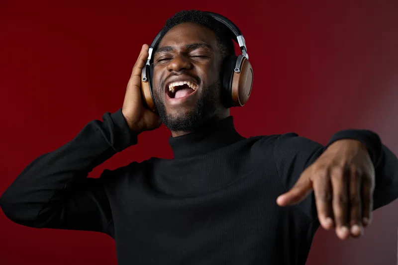 Happy young man enjoying music with headphones, expressing joy and energy in a vibrant red background while wearing a stylish black turtleneck