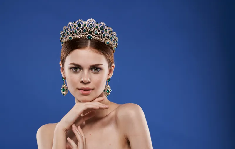 Привлекательная женщина с обнаженными плечами, короной на голове, украшениями и макияжем на синем фоне