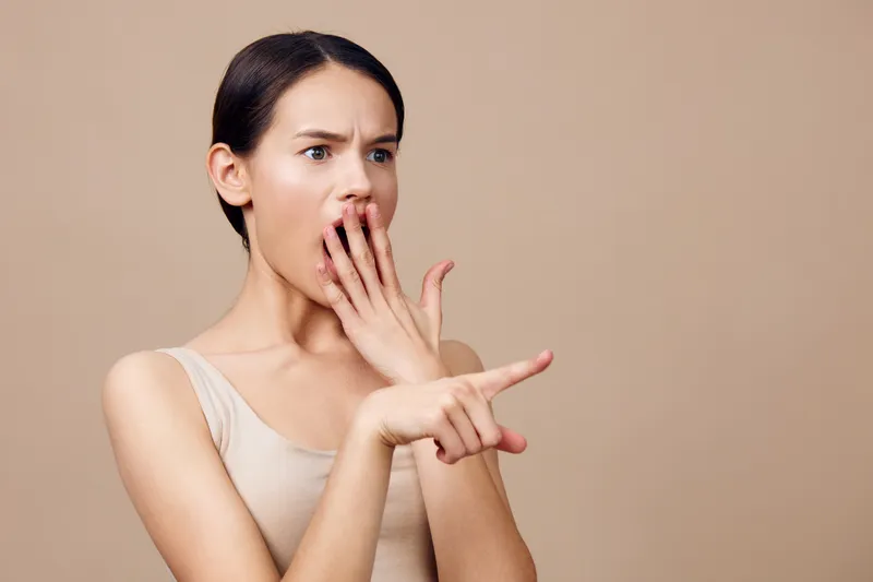 Surprised young woman with long dark hair in a nude top expressing shock on a plain beige background, ideal for emotional and reaction concepts