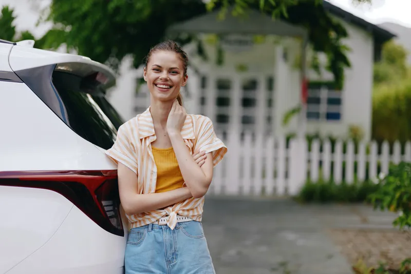 Весёлая молодая женщина стоит рядом с белым автомобилем и тепло улыбается