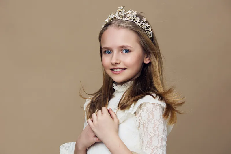Young girl with a crown, smiling joyfully, wearing a lace dress against a soft beige background, conveying innocence and elegance, ideal for beauty or child related concepts