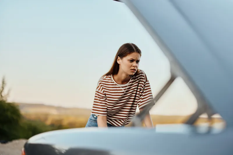 Молодая женщина-водитель в ужасе смотрит на автомобиль, открыв капот