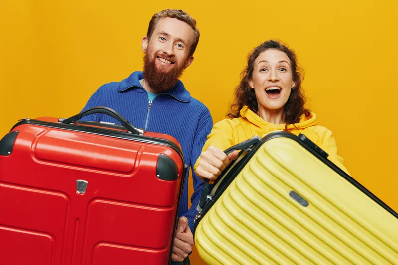 Мужчина и женщина улыбаются, держа в руках чемоданы, причем желтый и красный чемоданы стоят криво и весело