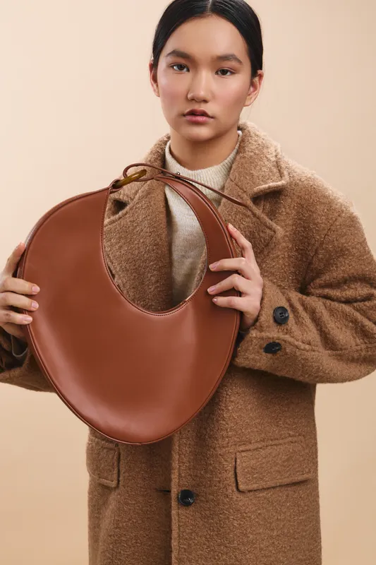 asian woman wearing a woolen coat holds a round brown handbag, posing for a fashion portrait with a neutral backdrop, conveying elegance and poised confidence.