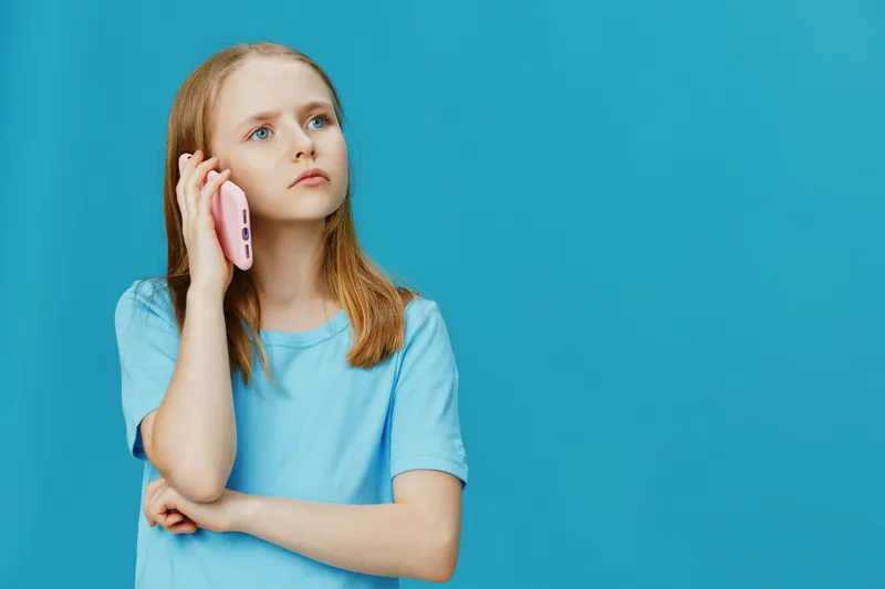 Thoughtful girl talking on a pink phone against a blue background, wearing a casual light blue t shirt, capturing a moment of curiosity and contemplation
