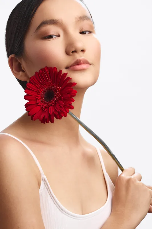 Asian woman holds a vibrant red flower near her face, posing in a studio with soft light. The calm expression and minimal attire emphasize beauty, artistry, and elegant simplicity in contemporary