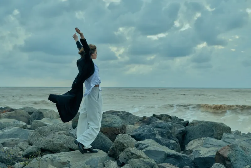 Woman standing on jagged rocks by the sea, wind sweeping her dress as she reaches upward, dramatic coast scene with overcast sky, outdoor moment filled with resilience and freedom