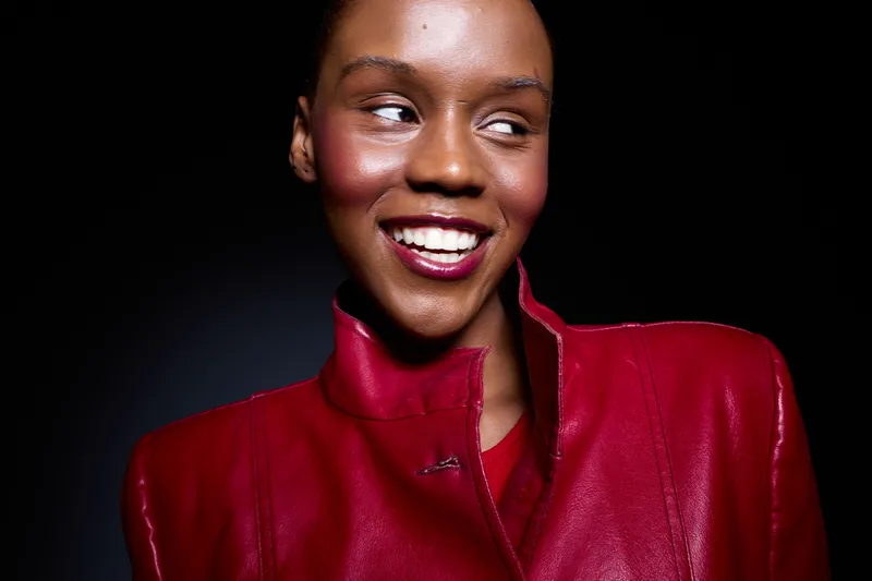 Smiling young black woman in red leather jacket looks sideways with happy expression. Studio portrait on dark background. Concept for fashion, beauty, confidence and positive emotions.
