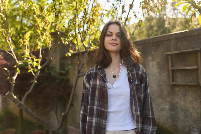 Casual woman lifestyle portrait outdoors wearing a plaid shirt over a white tee, a pendant necklace, and a relaxed smile in a sunlit garden setting in sunshine