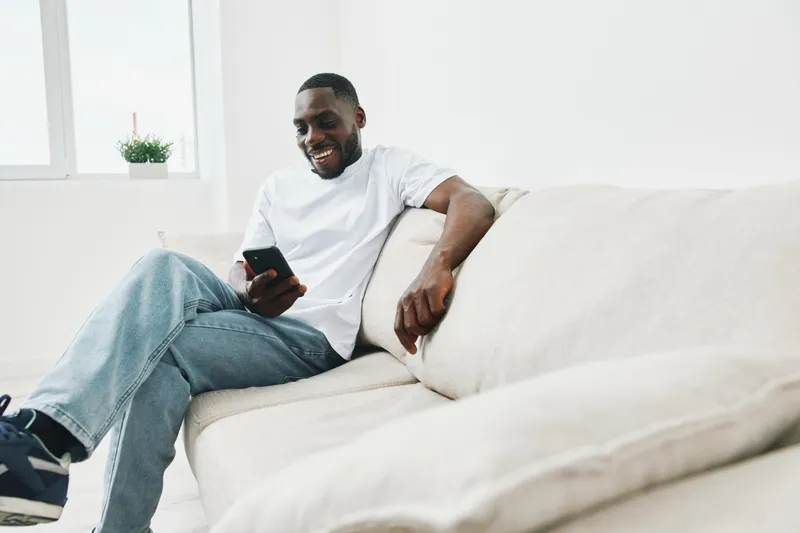 Relaxed African American man enjoying leisure time while using smartphone on a cozy couch in a bright, minimalistic living space, capturing joy and comfort