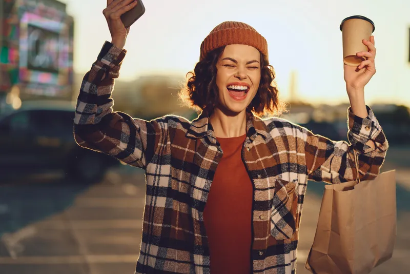 Woman with beanie holding coffee and takeaway bag, smiling with phone in city morning, shopping happiness and candid authenticity captured in golden hour glow, mindful living and emotional