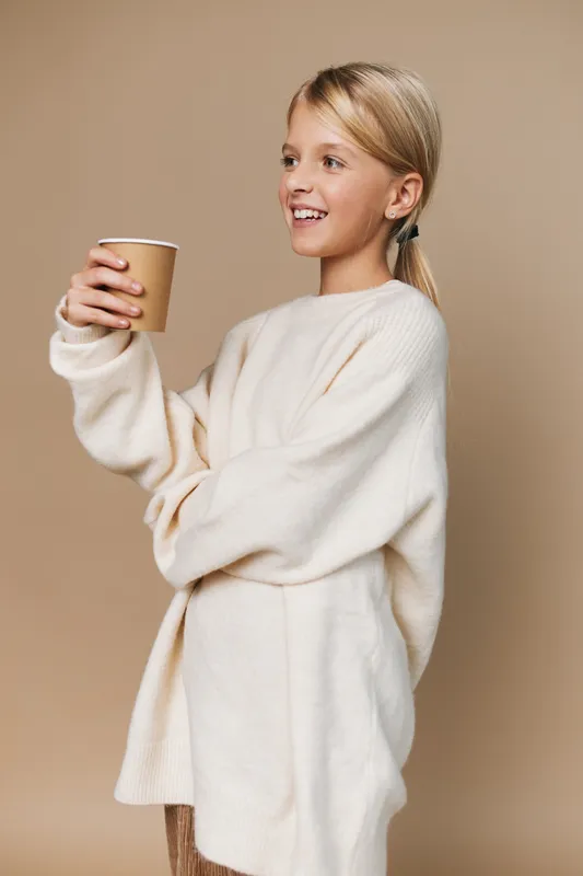 A cheerful girl in an oversized sweater holds a warm cup, smiling softly against a neutral beige background, conveying comfort, simplicity, and cozy everyday warmth.