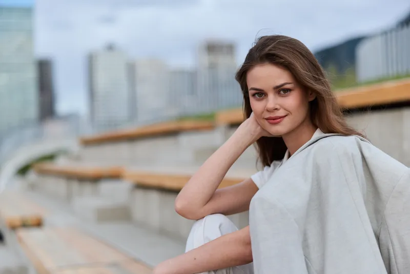Young woman with confident smile sitting on outdoor urban benches wearing light casual clothing in soft natural colors