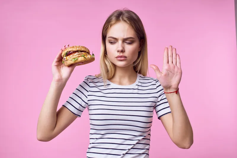 Весёлая женщина держит в руках фастфуд и перекусывает вкусной едой на розовом фоне
