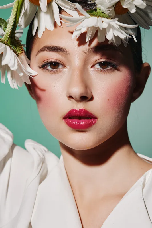 Close-up of a young woman with vivid pink lipstick and delicate flowers in her hair. She wears a white outfit against a soft pastel background, perfect for fashion and beauty.