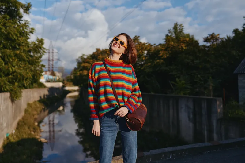 Woman in a colorful striped sweater and sunglasses smiling outdoors by an urban canal, wearing jeans and a leather crossbody bag, relaxed travel lifestyle, walking and enjoying sunny autumn day.
