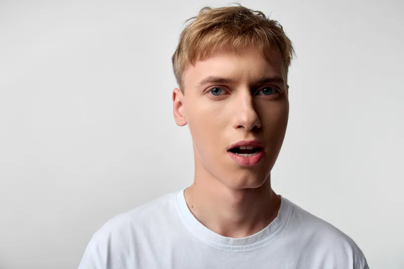 Surprised, young man, closeup, white t shirt, facial expression, astonishment captured in a studio setting with plain background, direct gaze, clear skin tone, authentic moment.