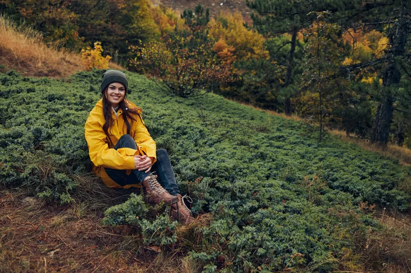 Молодая женщина в жёлтой куртке сидит на холме в лесу, наслаждаясь спокойным моментом среди красоты природы