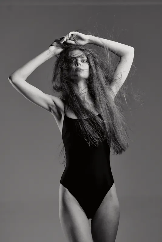 Black and white portrait of a woman in bodysuit posing with stretched arms and long hair, artistic and elegant monochrome fashion image.