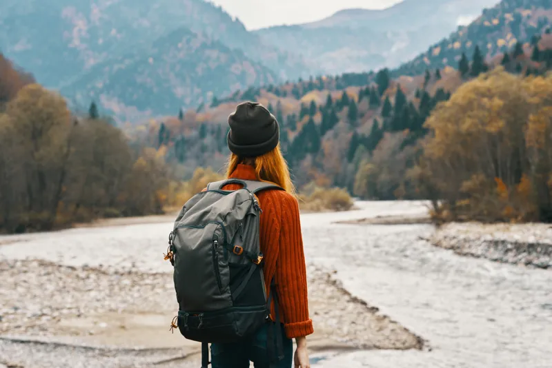 Женщина стоит на берегу реки и смотрит на горы вдалеке, пейзаж, осень, рюкзак, туризм, модель