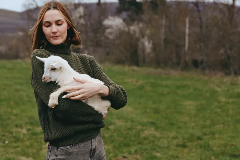 Calm woman outdoors holding a goat kid close, gentle expression, olive green sweater, soft natural light, rural field backdrop, relaxed posture, warm tones, candid lifestyle moment, fresh countryside