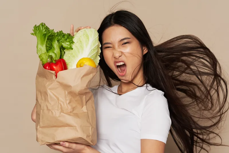 Эмоциональная азиатская женщина с продуктами в пакете на бежевом фоне гримасничает и выражает эмоции