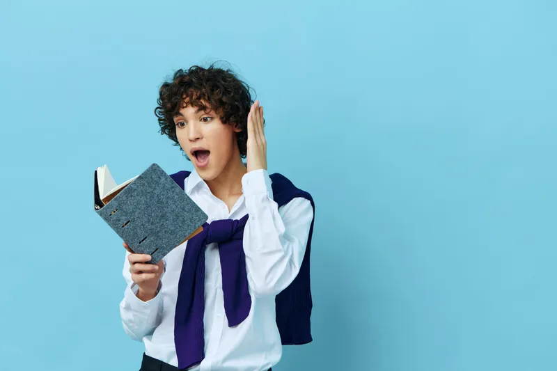 Кудрявый парень с книгой, в куртке, на изолированном фоне, в образе студента колледжа