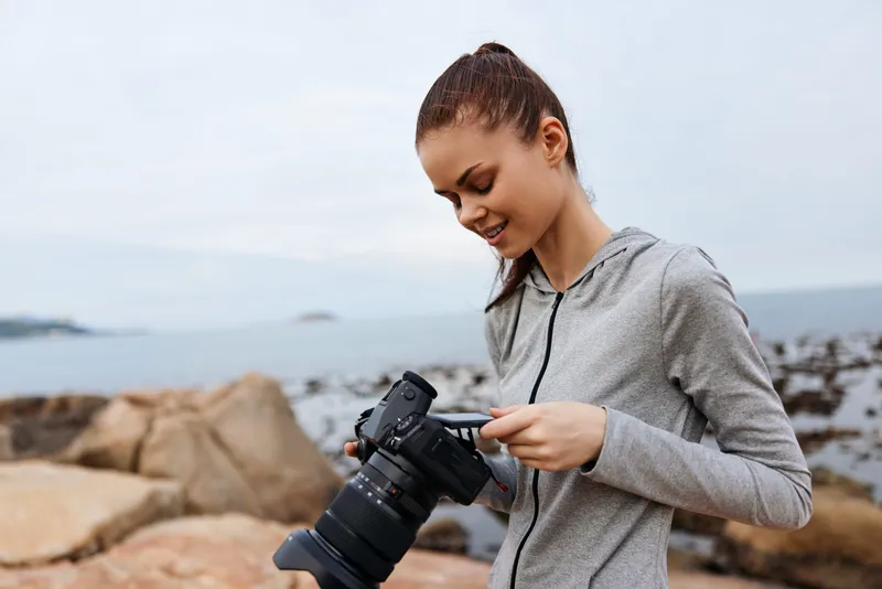 Young woman enjoying photography by the ocean, wearing a grey hoodie, smiling, capturing moments with a camera, nature background, spring or summer vibe