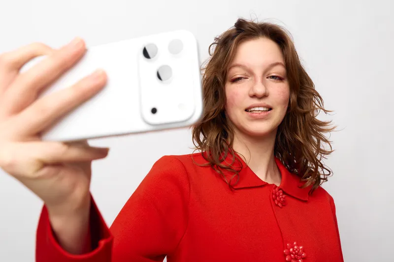 Young woman in red sweater takes selfie with white smartphone on white background. Casual style, natural expression, and modern technology concept for social media and communication.