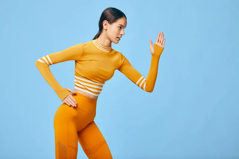 A fit woman in a bright orange athletic set posed in a dynamic running stance against a clean light blue studio background, emphasizing fitness, strength, posture and focused active lifestyle.