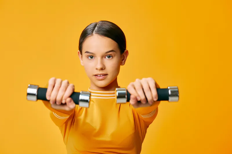 Young woman fitness portrait holding two dumbbells forward, wearing yellow sports top, focused expression, strength training and exercise concept in bright orange studio background for active