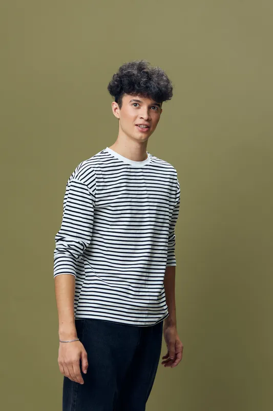 young man with curly hair wearing striped long sleeve shirt and dark pants standing against olive green background, casual style and relaxed expression