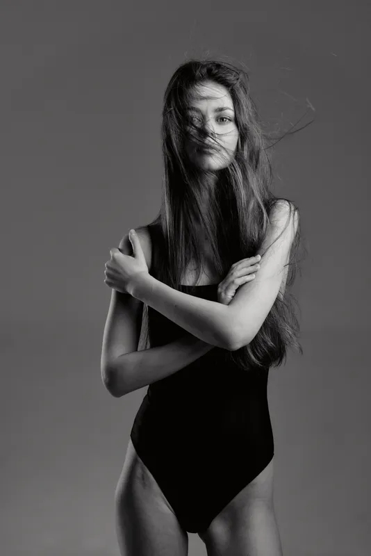 Black and white portrait of a young woman with long hair wearing a black bodysuit, expressing calm and confidence in studio lighting.