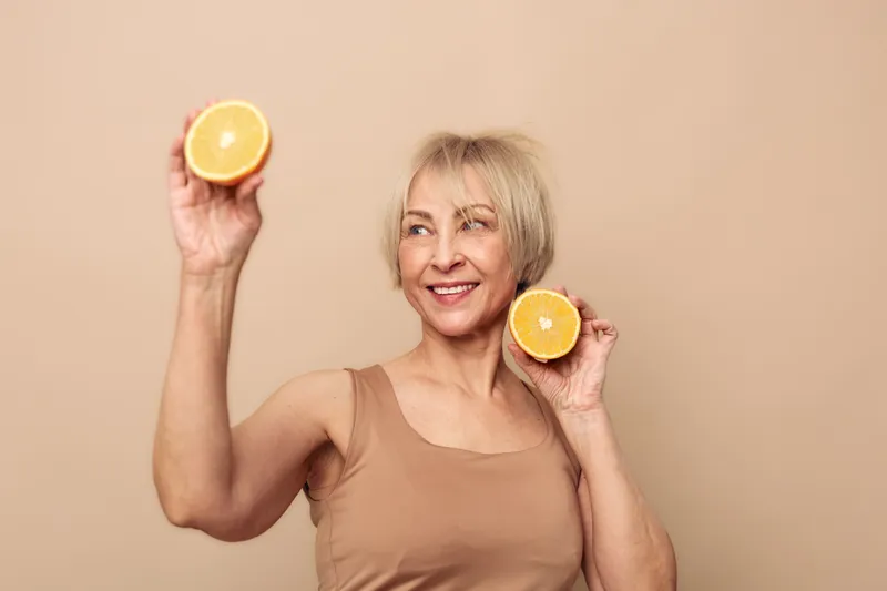 Smiling mature woman with short blonde hair holds two orange halves near her face, wearing beige sleeveless top. Concept for healthy lifestyle, nutrition, vitamin C, and wellness.