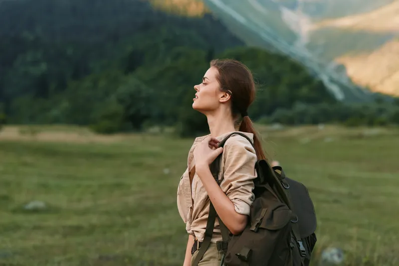 A woman with a backpack stands in a wide meadow, gazing upward as breeze lifts her hair. The scene conveys adventure, calm, and a strong connection with nature.