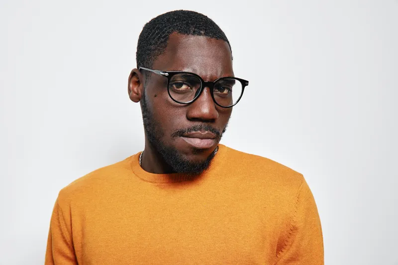 African American man with dark skin wearing glasses and orange sweater looking at camera with serious expression on white background. Portrait, fashion and style concept.