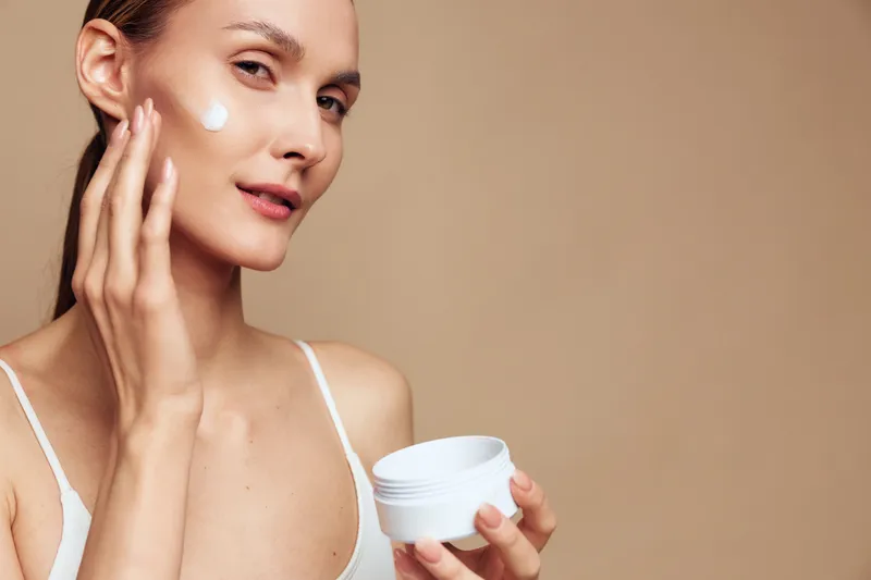 Young woman applying moisturizing cream on face with fingers, clear skin, neutral background, skincare routine, natural beauty, soft lighting, close-up portrait