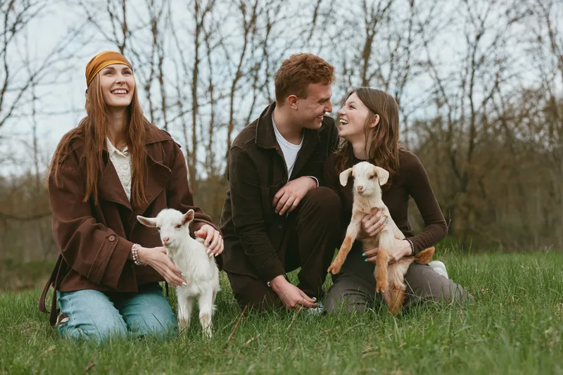 Lifestyle outdoors friends, three smiling people in casual clothes with a goat kid, playful and warm mood, sitting on grass in a park, natural candid moment of friendship