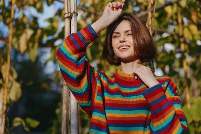 Woman in a colorful striped sweater smiling outdoors, bob haircut, leaning on a wooden pole in a sunlit garden. Casual outfit, cheerful young adult, lifestyle fashion portrait.