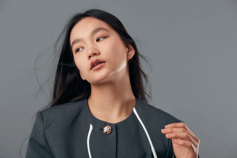 Woman with long dark hair posing in gray business jacket in studio with neutral background, looking sideways with natural makeup and serious facial expression for portrait photography.
