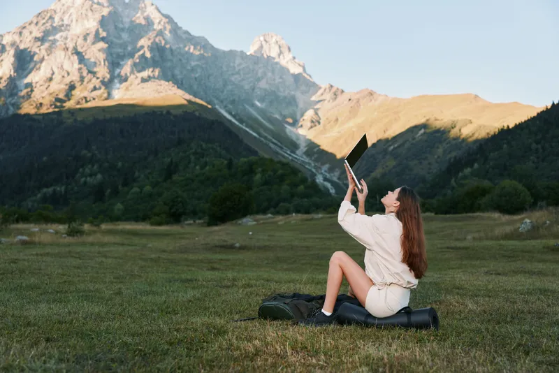 A cheerful traveler sits on a grassy meadow, raising a laptop toward the bright sky to capture a photo, with rugged mountains behind, creating a joyful outdoor vibe.